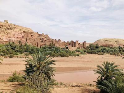 A wide landscape shot of a large, reddish-brown earthen fortified village (ksar) built into a hillside, with a dry riverbed and palm trees in the foreground.