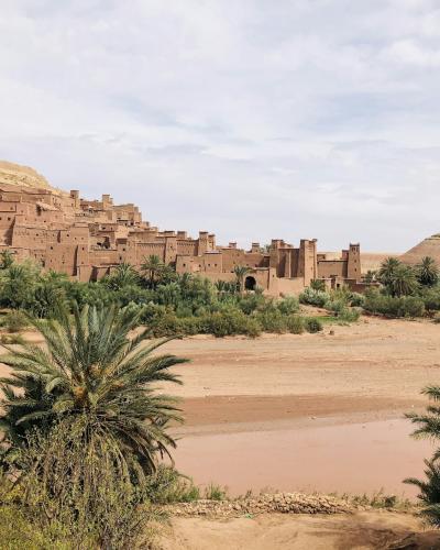 A wide landscape shot of a large, reddish-brown earthen fortified village (ksar) built into a hillside, with a dry riverbed and palm trees in the foreground.