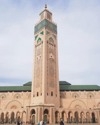 A large sandstone mosque with a tall, square minaret featuring green and white tilework, set against a cloudy sky with people at the base.
