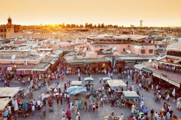A high-angle view of a crowded public square at sunset, featuring numerous market stalls with awnings, dense crowds of people, and surrounding buildings with flat roofs and a prominent minaret on the left.