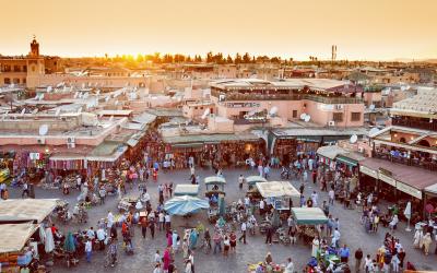 A high-angle view of a crowded public square at sunset, featuring numerous market stalls with awnings, dense crowds of people, and surrounding buildings with flat roofs and a prominent minaret on the left.