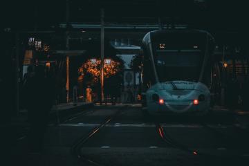 A white high-speed train (Al Boraq) is stopped at a station platform at night, with its headlights on and a person standing on the left side of the tracks.