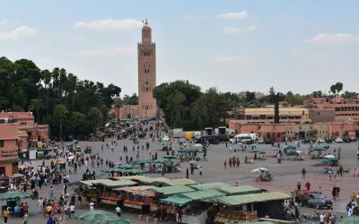 A large, open public square filled with people, market stalls with green umbrellas, and a tall, distinctive minaret tower in the background under a partly cloudy sky.