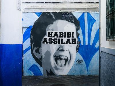A black and white mural of a laughing child's face with the text "HABIBI ASSILAH" painted on a white wall next to a blue-painted section and a barred window.