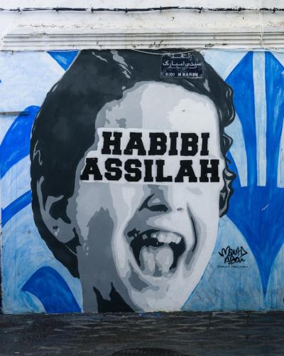 A black and white mural of a laughing child's face with the text "HABIBI ASSILAH" painted on a white wall next to a blue-painted section and a barred window.
