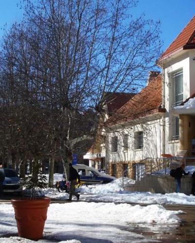 A white building with a red-tiled roof and snow on its roof, set against a clear blue sky, with bare trees and people walking along a snowy street.
