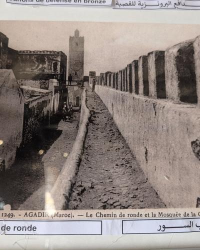 A vintage black-and-white photograph showing a narrow, elevated stone walkway (chemin de ronde) along a fortified wall, with a minaret visible in the background and people walking along the path.