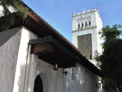 A low-angle shot of a white building with Moorish architectural features, including a horseshoe archway, a tiled roof, and a square tower with a crenellated top and a flagpole, set against a blue sky with trees.