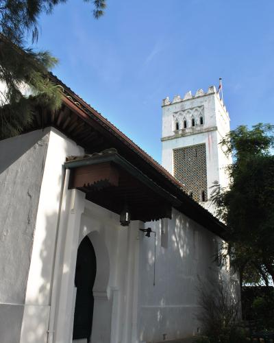 A low-angle shot of a white building with Moorish architectural features, including a horseshoe archway, a tiled roof, and a square tower with a crenellated top and a flagpole, set against a blue sky with trees.
