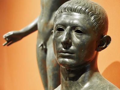 A close-up photograph of a bronze bust of a young man with short wavy hair, with another bronze statue visible in the blurred background against an orange wall.