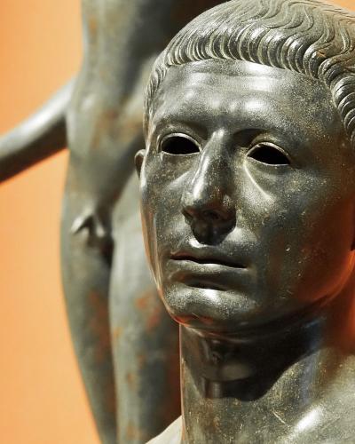 A close-up photograph of a bronze bust of a young man with short wavy hair, with another bronze statue visible in the blurred background against an orange wall.