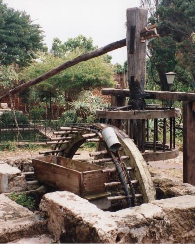 A large wooden noria (waterwheel) set within a stone basin in a garden with trees and flowers, with people walking in the background.