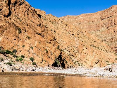 A wide shot of a river flowing through a steep, layered canyon with reddish-brown rock walls and sparse green vegetation along the banks under a clear blue sky.