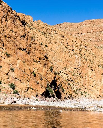 A wide shot of a river flowing through a steep, layered canyon with reddish-brown rock walls and sparse green vegetation along the banks under a clear blue sky.