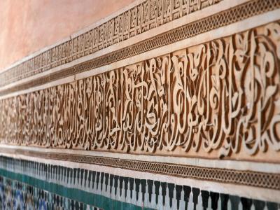 A close-up view of intricately carved stucco walls featuring Arabic calligraphy and geometric patterns, situated above a band of green and white zellige tilework.