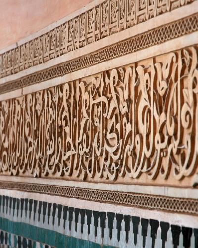 A close-up view of intricately carved stucco walls featuring Arabic calligraphy and geometric patterns, situated above a band of green and white zellige tilework.