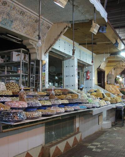A view inside a covered market alleyway featuring a confectionery stall displaying various Moroccan sweets and pastries, with tiled arches and hanging lights.
