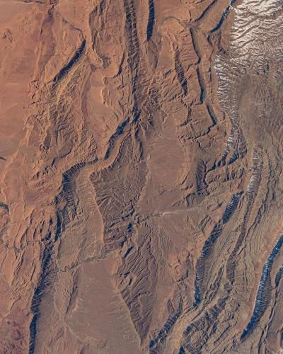 A high-altitude satellite or space station photograph showing a vast, arid landscape with distinct geological formations, including reddish desert plains on the left and rugged, snow-capped mountain ranges on the right.