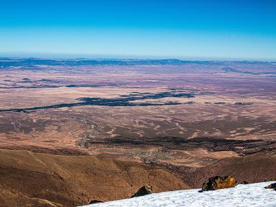 A high-altitude panoramic view looking out over a vast, arid desert plain from a snowy mountain ridge, with distant mountain ranges on the horizon under a clear blue sky.