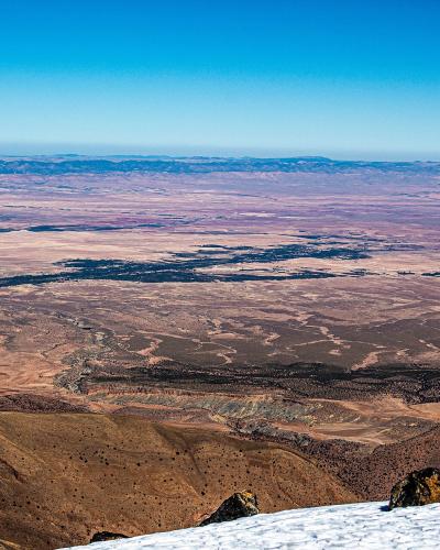 A high-altitude panoramic view looking out over a vast, arid desert plain from a snowy mountain ridge, with distant mountain ranges on the horizon under a clear blue sky.
