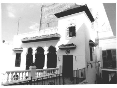 A black and white photograph of a white building featuring Moorish-style horseshoe arches, a tiled roof, and a balcony with a balustrade.