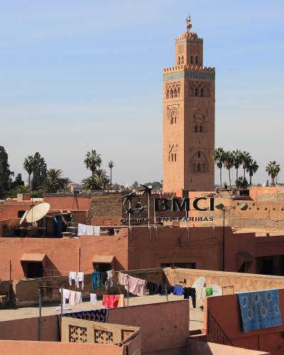 A tall, square, reddish-brown minaret with intricate geometric carvings and a decorative top rises above a dense urban landscape of terracotta-colored buildings, palm trees, and rooftop laundry under a clear blue sky.