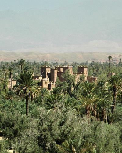 A large, traditional earthen kasbah with crenellated towers is visible rising above a dense grove of palm trees and other vegetation, with arid hills in the background.