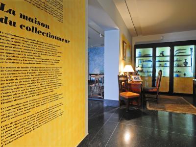 An interior view of a museum room featuring a large yellow wall panel with French text titled "La maison du collectionneur" and glass display cabinets containing decorative arts.