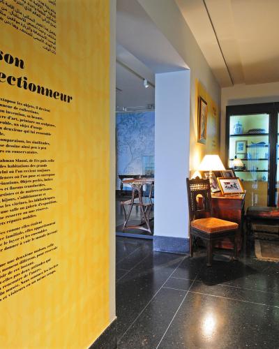 An interior view of a museum room featuring a large yellow wall panel with French text titled "La maison du collectionneur" and glass display cabinets containing decorative arts.