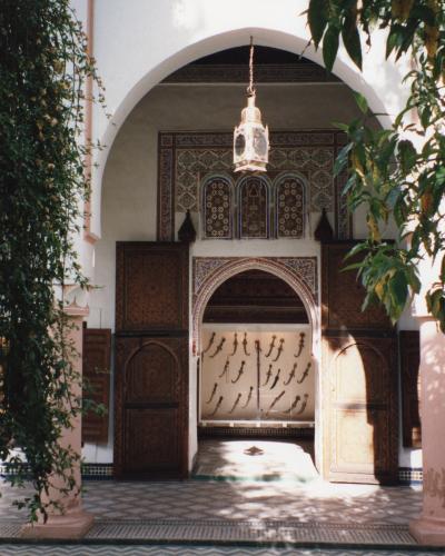 A view into a courtyard of a Moroccan riad or museum, featuring an ornate horseshoe archway with intricate tilework (zellij) and carved wood, leading to a room displaying a collection of daggers on the wall.