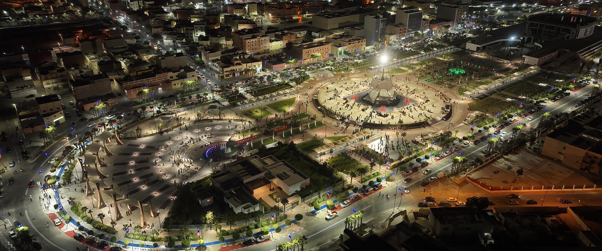 Aerial night view of a large, illuminated urban plaza with a central fountain, surrounded by curved roads, palm trees, and city buildings.
