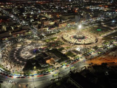 Aerial night view of a large, illuminated urban plaza with a central fountain, surrounded by curved roads, palm trees, and city buildings.