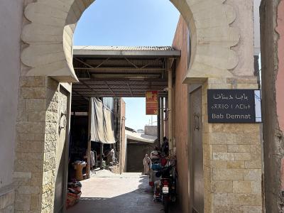A view through a scalloped stone archway into a narrow market alleyway, with a black sign on the right pillar reading "Bab Demnat" in Arabic, Tifinagh, and Latin script.