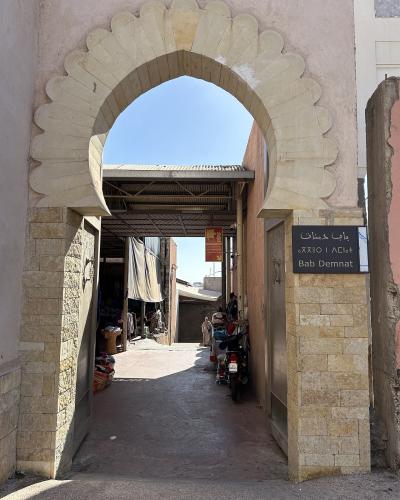A view through a scalloped stone archway into a narrow market alleyway, with a black sign on the right pillar reading "Bab Demnat" in Arabic, Tifinagh, and Latin script.
