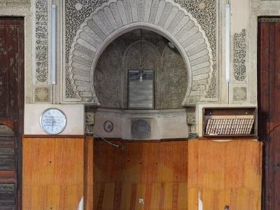 The image shows the prayer hall of a historic Islamic building featuring ornate stucco arches, marble columns, zellige tilework, and a mihrab niche, with wooden paneling and prayer mats on the floor.
