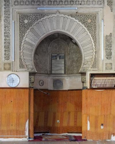 The image shows the prayer hall of a historic Islamic building featuring ornate stucco arches, marble columns, zellige tilework, and a mihrab niche, with wooden paneling and prayer mats on the floor.