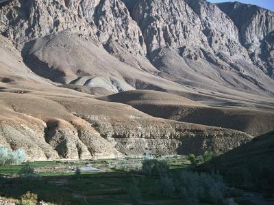 A landscape photograph showing a dry, arid valley with steep, rocky mountains in the background and a strip of green vegetation and trees along a riverbed in the foreground.
