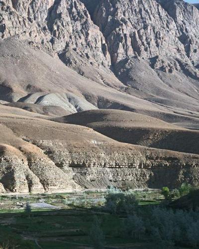 A landscape photograph showing a dry, arid valley with steep, rocky mountains in the background and a strip of green vegetation and trees along a riverbed in the foreground.