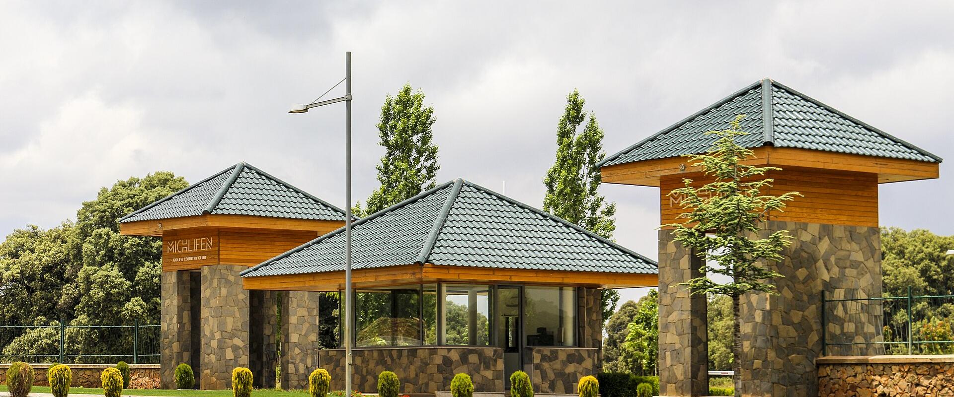 The image shows the entrance gate structures of a facility, featuring stone pillars, wooden accents, and green tiled roofs, with the name "MICHLIFEN" visible on the left structure.