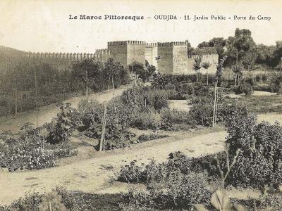 A vintage postcard showing a landscaped garden with young trees and flowerbeds in the foreground, backed by a large fortified wall with crenellations and a gate structure.