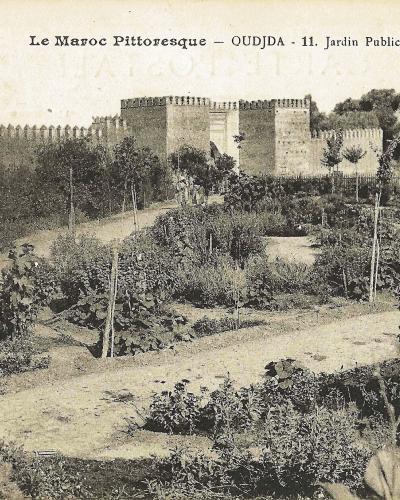 A vintage postcard showing a landscaped garden with young trees and flowerbeds in the foreground, backed by a large fortified wall with crenellations and a gate structure.
