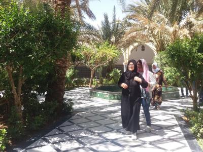 A paved garden path with a black and white geometric tile pattern, flanked by green trees and palm trees, with three women walking towards the camera and a small tiled fountain or pool in the background.