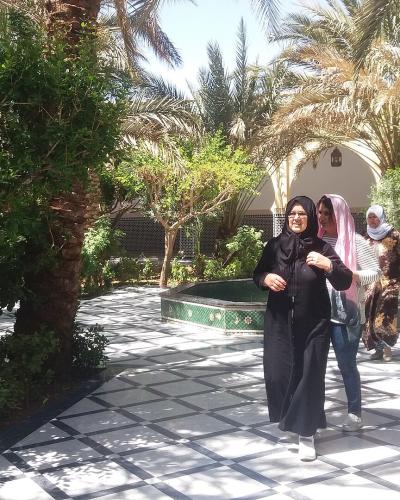 A paved garden path with a black and white geometric tile pattern, flanked by green trees and palm trees, with three women walking towards the camera and a small tiled fountain or pool in the background.
