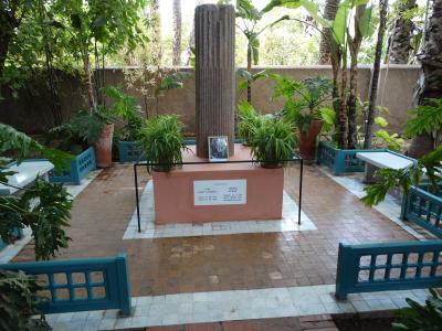A memorial site featuring a central stone pillar and a plaque dedicated to Yves Saint Laurent and Pierre Bergé, surrounded by teal-colored benches and lush tropical plants.