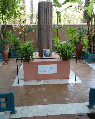 A memorial site featuring a central stone pillar and a plaque dedicated to Yves Saint Laurent and Pierre Bergé, surrounded by teal-colored benches and lush tropical plants.