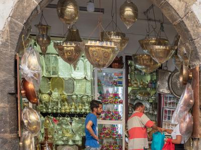 A souvenir shop displaying brass lanterns, trays, and tea sets viewed through a stone archway, with customers browsing inside.