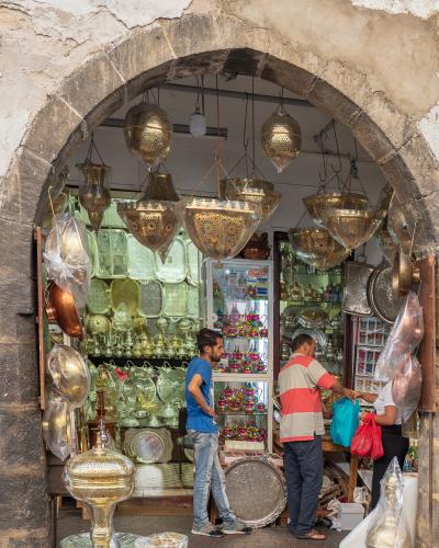 A souvenir shop displaying brass lanterns, trays, and tea sets viewed through a stone archway, with customers browsing inside.