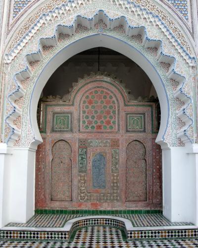 A courtyard with white walls, horseshoe arches, a central fountain, and a distinctive green-tiled roof structure in the background.