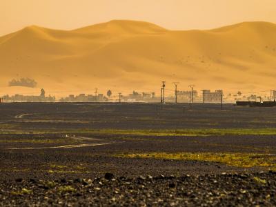 A landscape photograph showing a dark, rocky desert plain in the foreground with tire tracks, a line of buildings and utility poles in the mid-ground, and massive golden sand dunes rising in the background under a hazy sky.