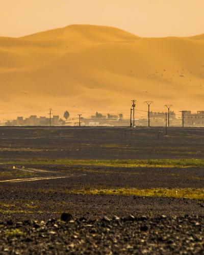 A landscape photograph showing a dark, rocky desert plain in the foreground with tire tracks, a line of buildings and utility poles in the mid-ground, and massive golden sand dunes rising in the background under a hazy sky.
